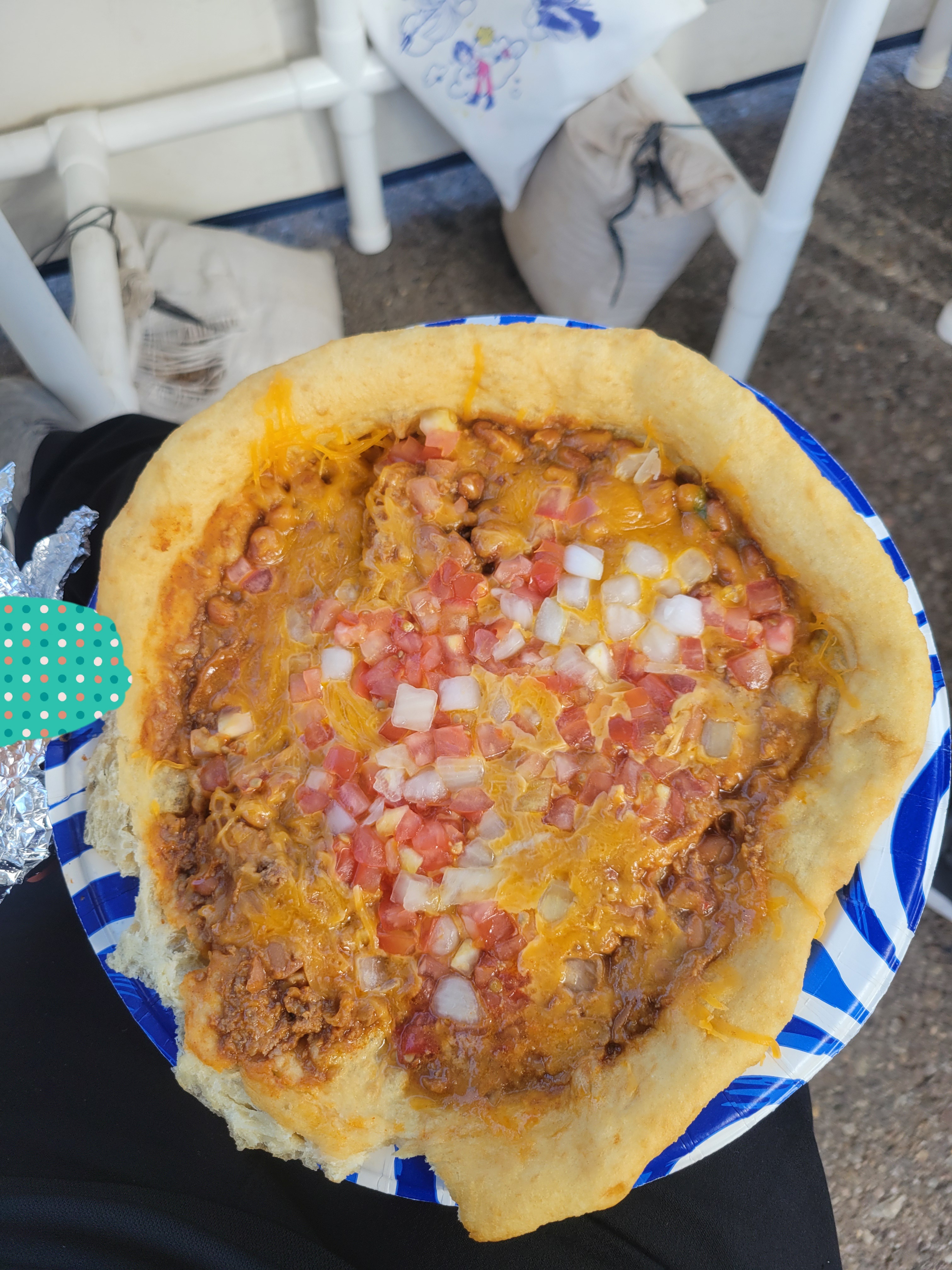Hand holds plate of fried bread topped with meat, cheese, beans, tomatoes, and onions.